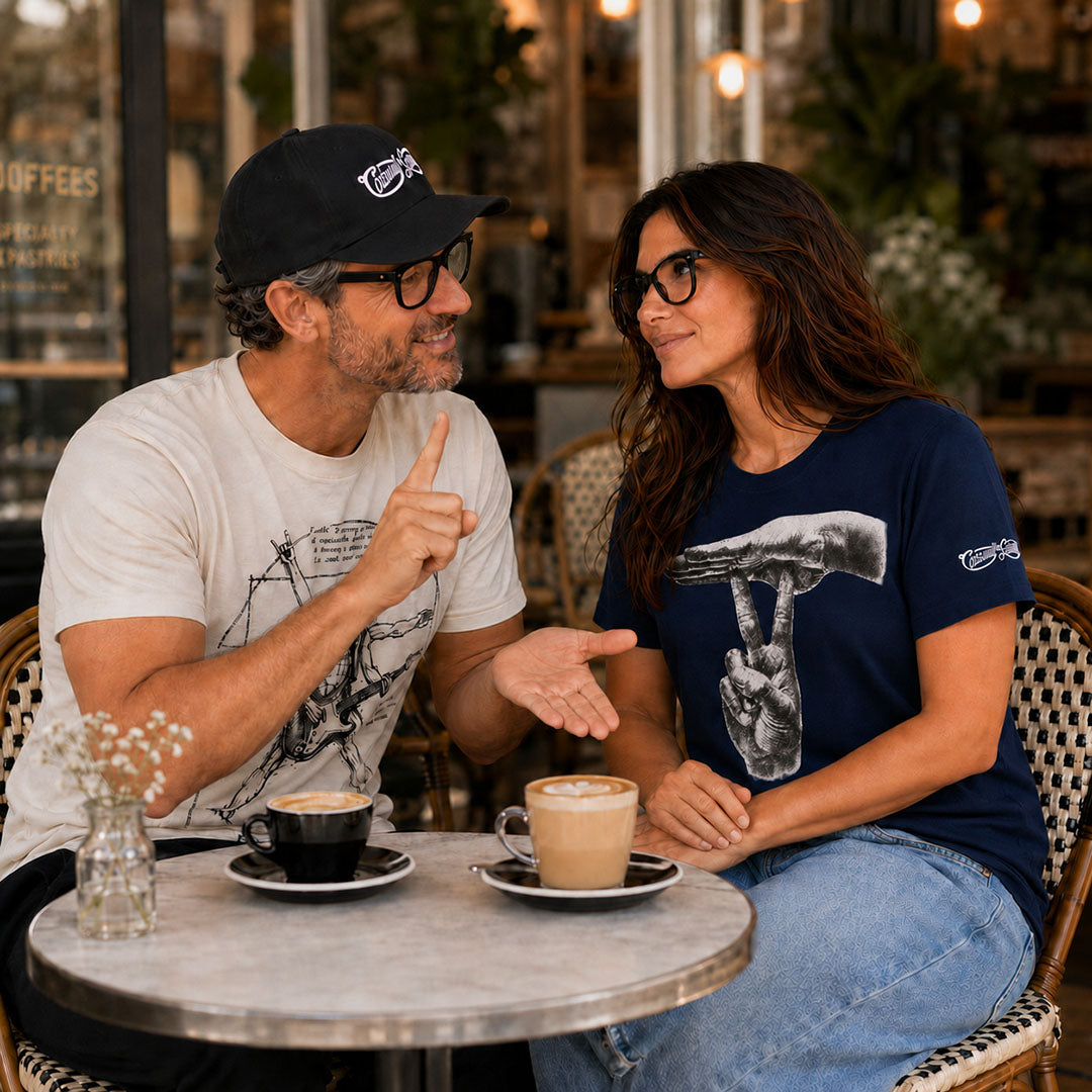 A man and a woman sitting at a cafe table, both wearing graphic t-shirts. The man wears a white graphic tee and a black cap, while the woman wears a navy blue graphic tee with a graphic of the ASL pun "UNDER-STAND."
