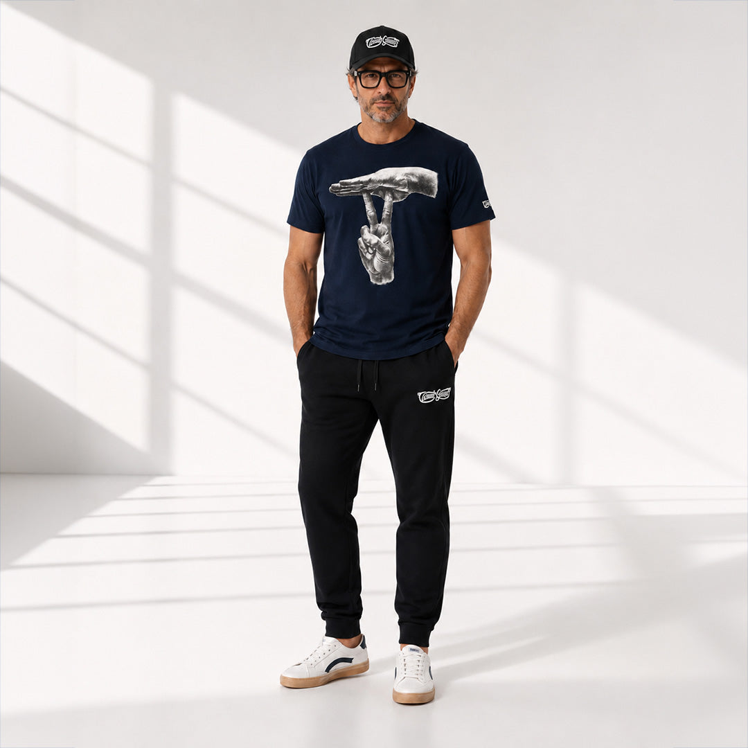 A man modeling a navy blue t-shirt with a large hammer graphic, black sweatpants, and white sneakers in a bright studio with window-like shadows.