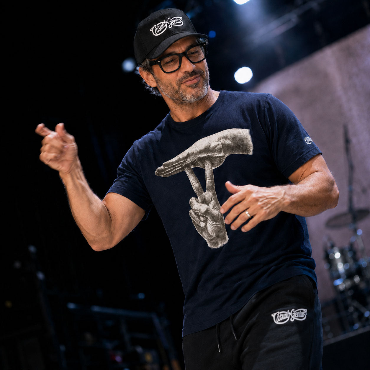 A man signing on a stage wearing a navy blue t-shirt with a large grayscale graphic of two hands forming the pun UNDER-STAND in ASL, a black baseball cap with a white logo, and black pants.