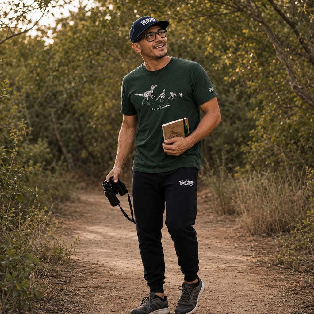 Man birdwatching in a forest, holding binoculars and an ornithology journal, wearing a green t-shirt and black joggers.