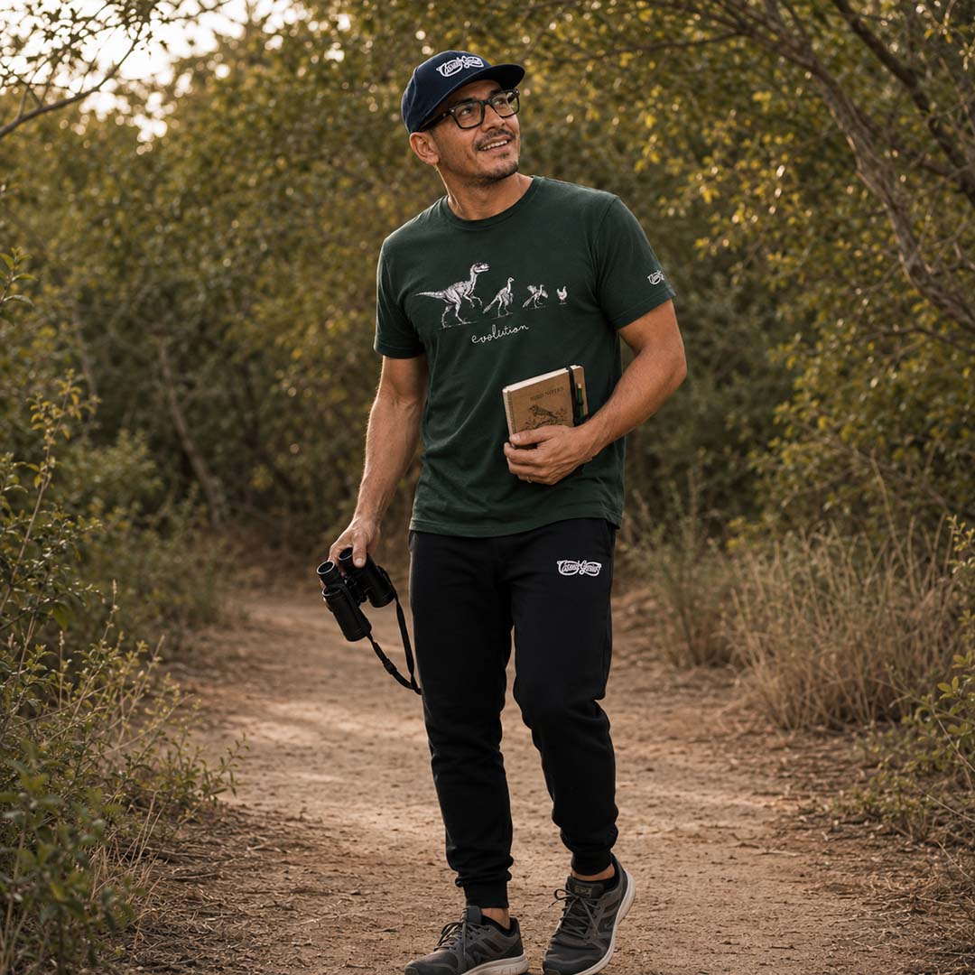 Man birdwatching in a forest, holding binoculars and an ornithology journal, wearing a green t-shirt and black joggers.