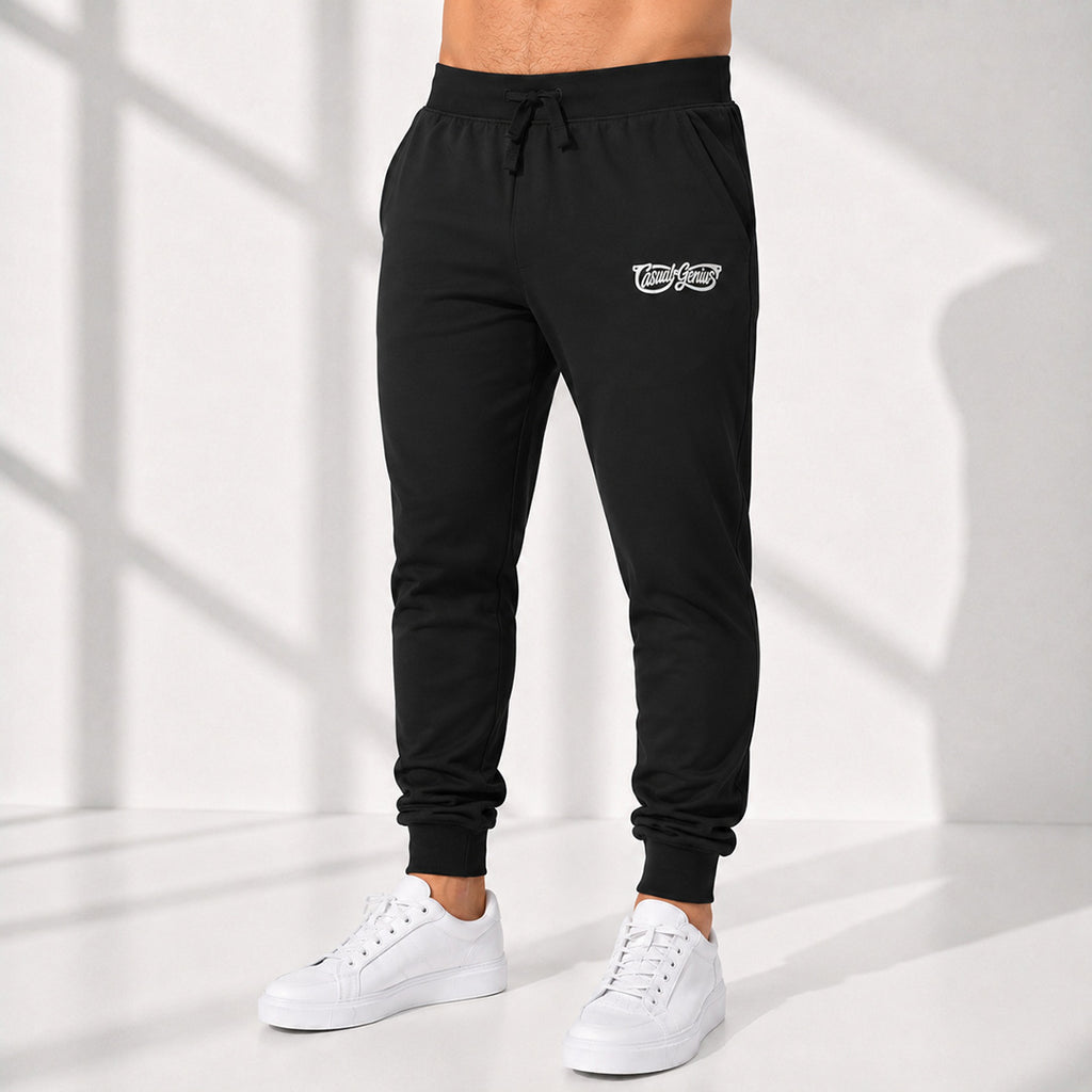 A person wearing black jogger sweatpants with a small white Casual Genius graphic logo on the upper left thigh, paired with white sneakers, standing in a bright studio with window-shadow lighting.