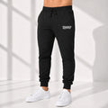 A person wearing black jogger sweatpants with a small white Casual Genius graphic logo on the upper left thigh, paired with white sneakers, standing in a bright studio with window-shadow lighting.