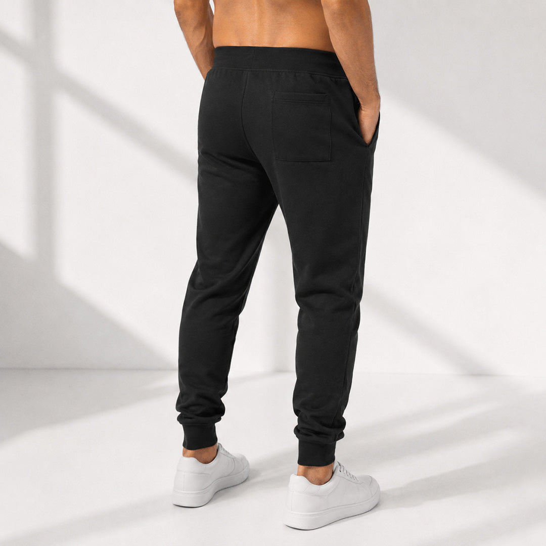 Black jogger sweatpants with an elastic waistband and ankle cuffs, shown from the back on a model wearing white sneakers against a white background with soft window shadows.