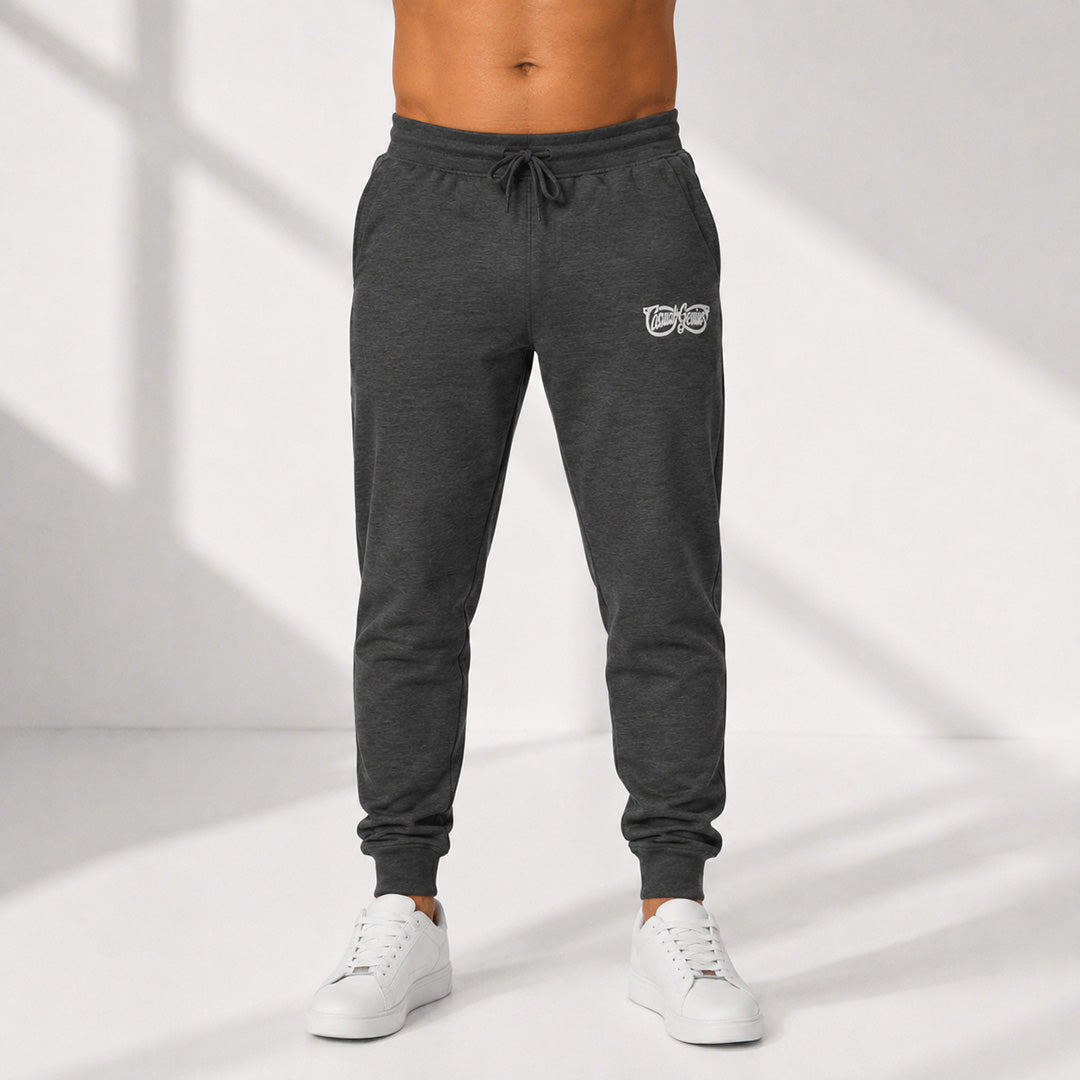 A person wearing dark heather gray jogger sweatpants with a small white Casual Genius graphic logo on the left thigh, paired with white sneakers, standing against a white wall with soft diagonal shadows.