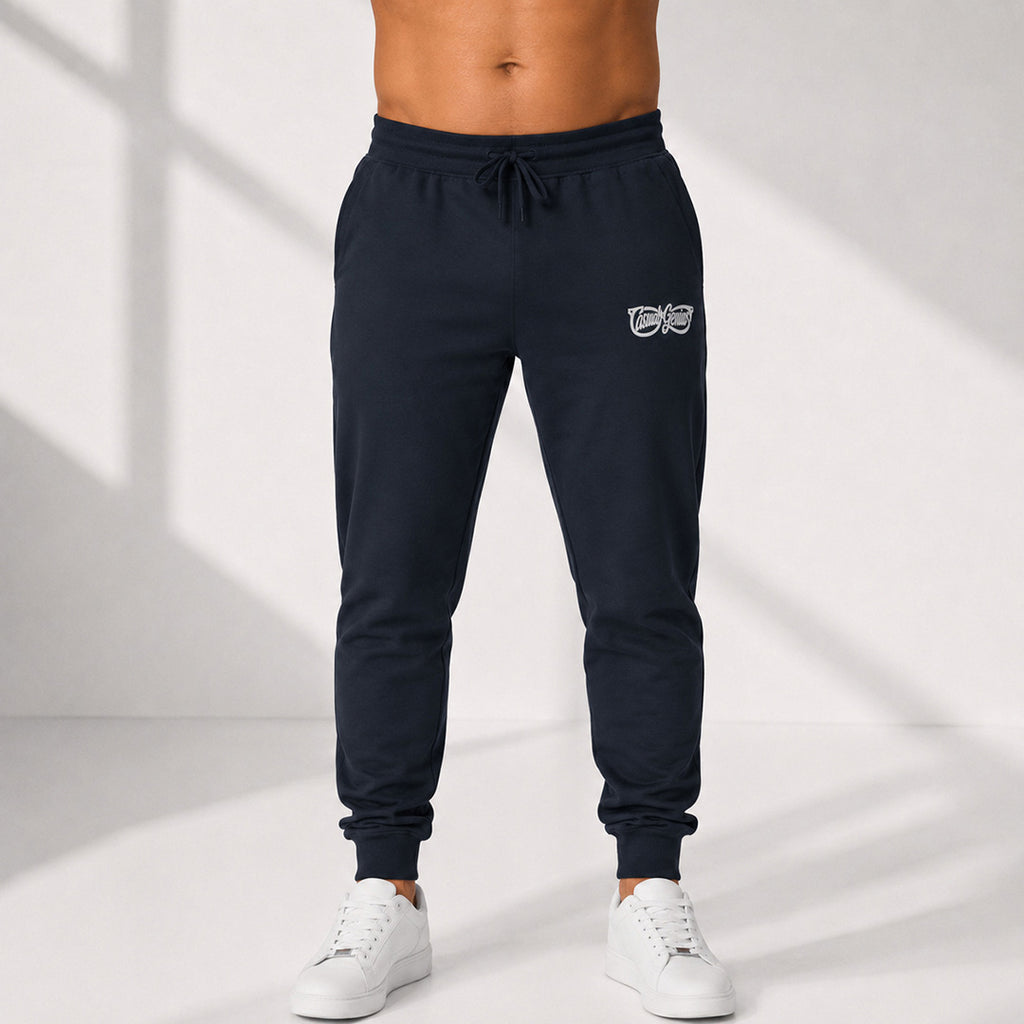 A person wearing dark navy jogger sweatpants with a small white Casual Genius graphic on the upper left thigh, paired with white sneakers, shown from the waist down against a light studio background with soft window shadows.