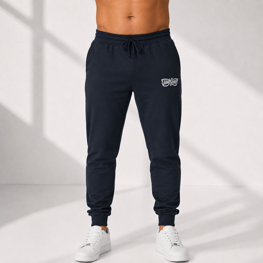 A person wearing dark navy jogger sweatpants with a small white Casual Genius graphic on the upper left thigh, paired with white sneakers, shown from the waist down against a light studio background with soft window shadows.