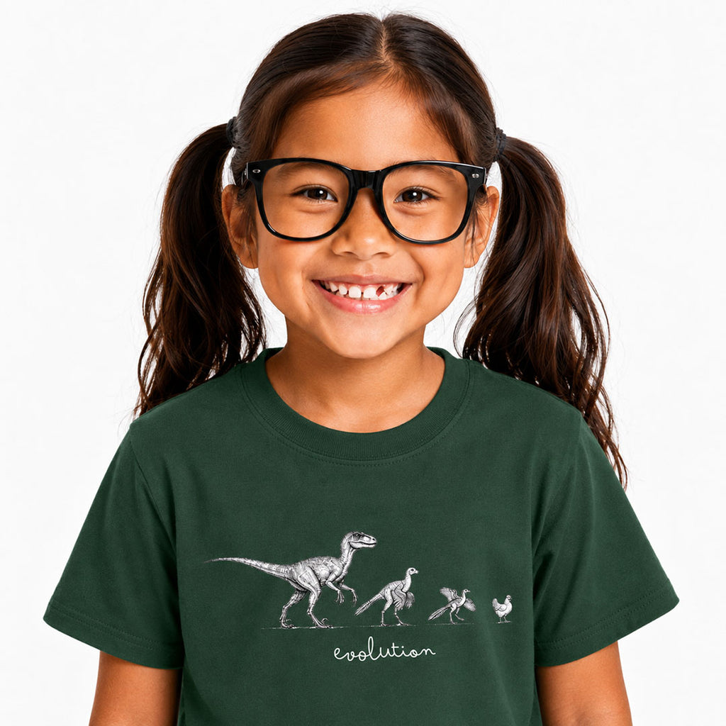 A young girl with dark hair in pigtails and black glasses is smiling, wearing a dark green t-shirt with a white graphic showing dinosaur evolution and the word 'evolution' written below.