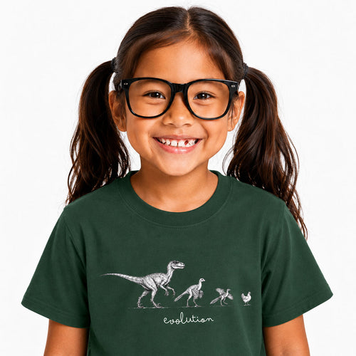 A young girl with dark hair in pigtails and black glasses is smiling, wearing a dark green t-shirt with a white graphic showing dinosaur evolution and the word 'evolution' written below.