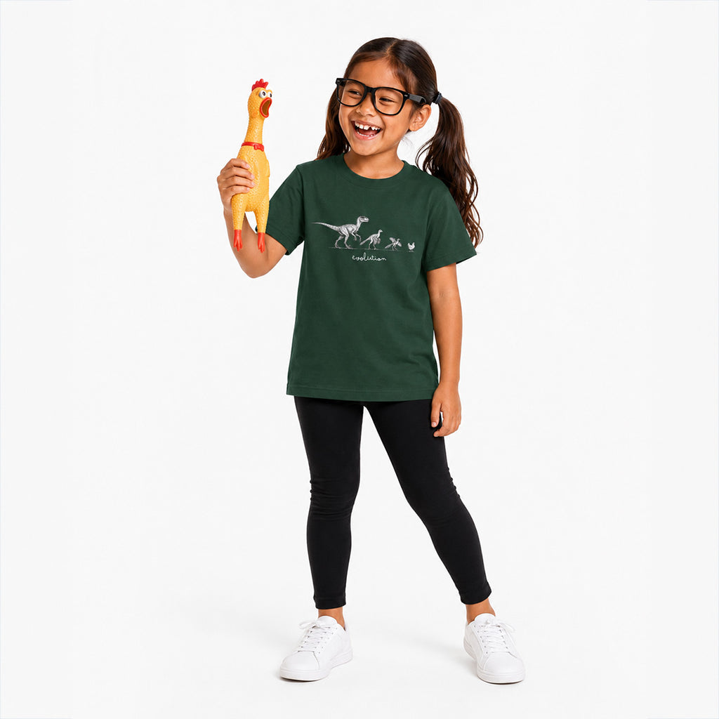 A young girl with glasses and pigtails stands against a white studio background, smiling and holding a yellow rubber chicken toy. She is wearing a dark green t-shirt with a white dinosaur graphic and text, black leggings, and white sneakers.