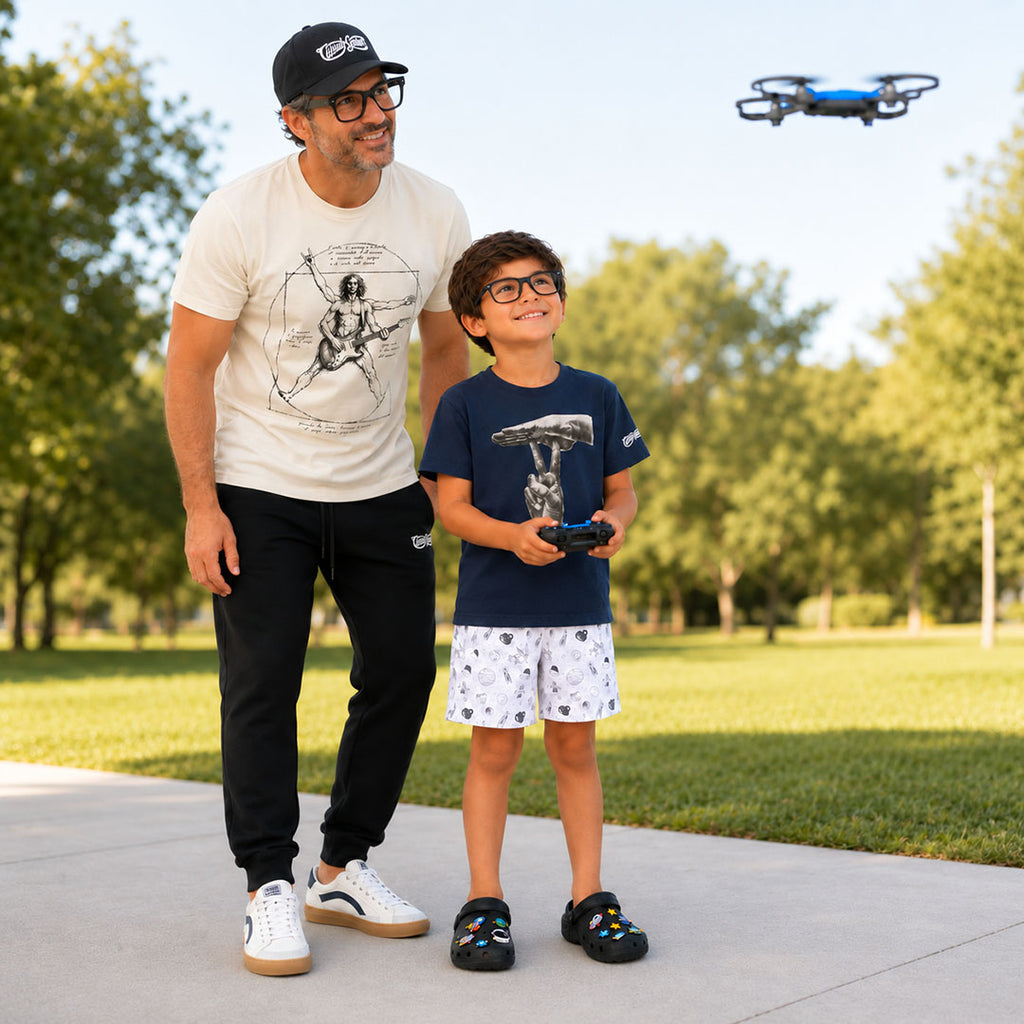 A father and son outdoors in a park, with the boy holding a remote control for a small blue drone flying overhead. The man wears a white graphic t-shirt, black sweatpants, and white sneakers, while the boy wears a navy t-shirt with a graphic of the ASL pun "UNDER-STAND", white shorts, and black sandals.