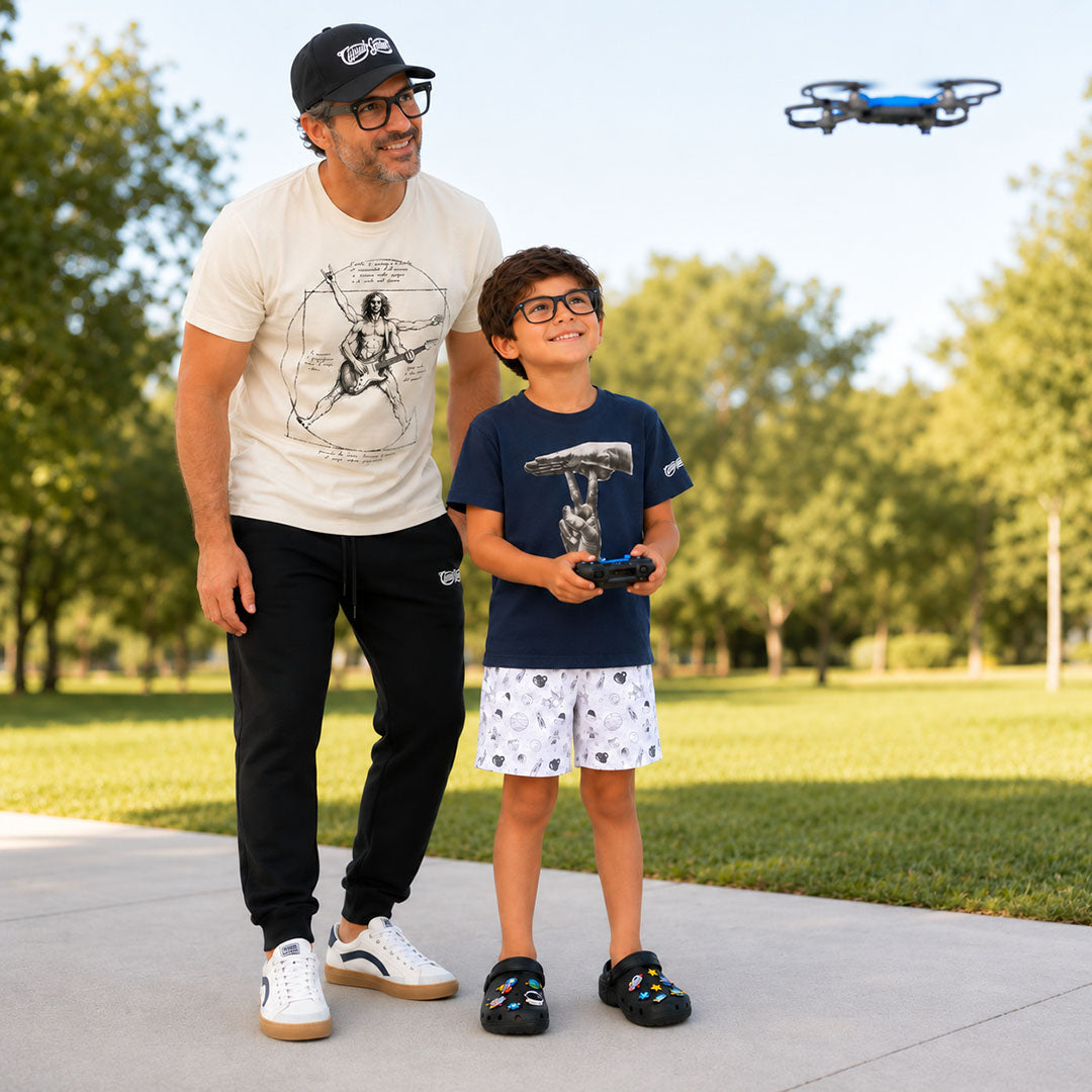 A father and son outdoors in a park, with the boy holding a remote control for a small blue drone flying overhead. The man wears a white graphic t-shirt, black sweatpants, and white sneakers, while the boy wears a navy t-shirt with a graphic of the ASL pun "UNDER-STAND", white shorts, and black sandals.