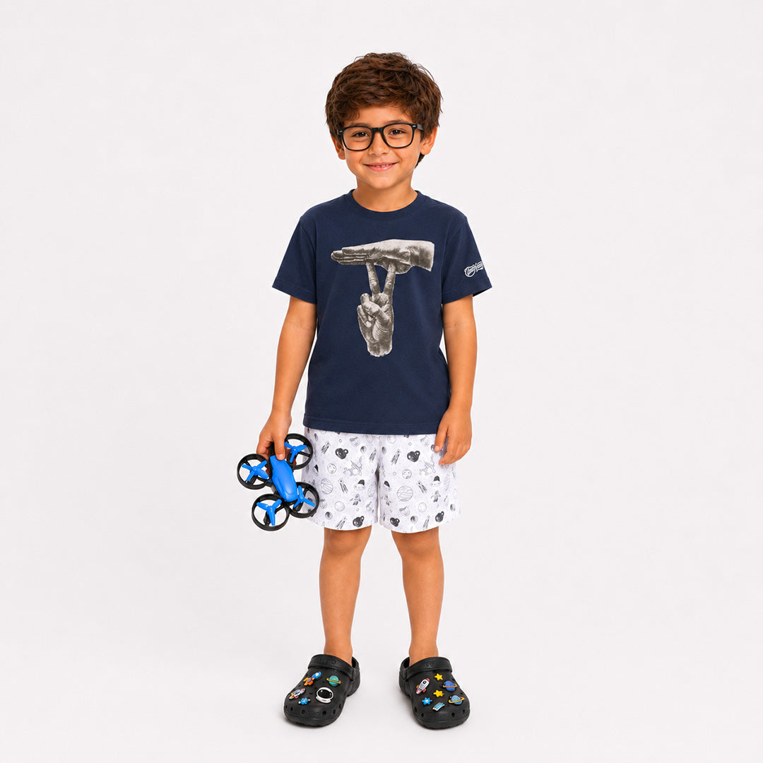 A young boy wearing a navy blue t-shirt with a hammer graphic, white shorts with a black pattern, and black clogs, holding a blue and black remote-controlled drone against a light gray background.