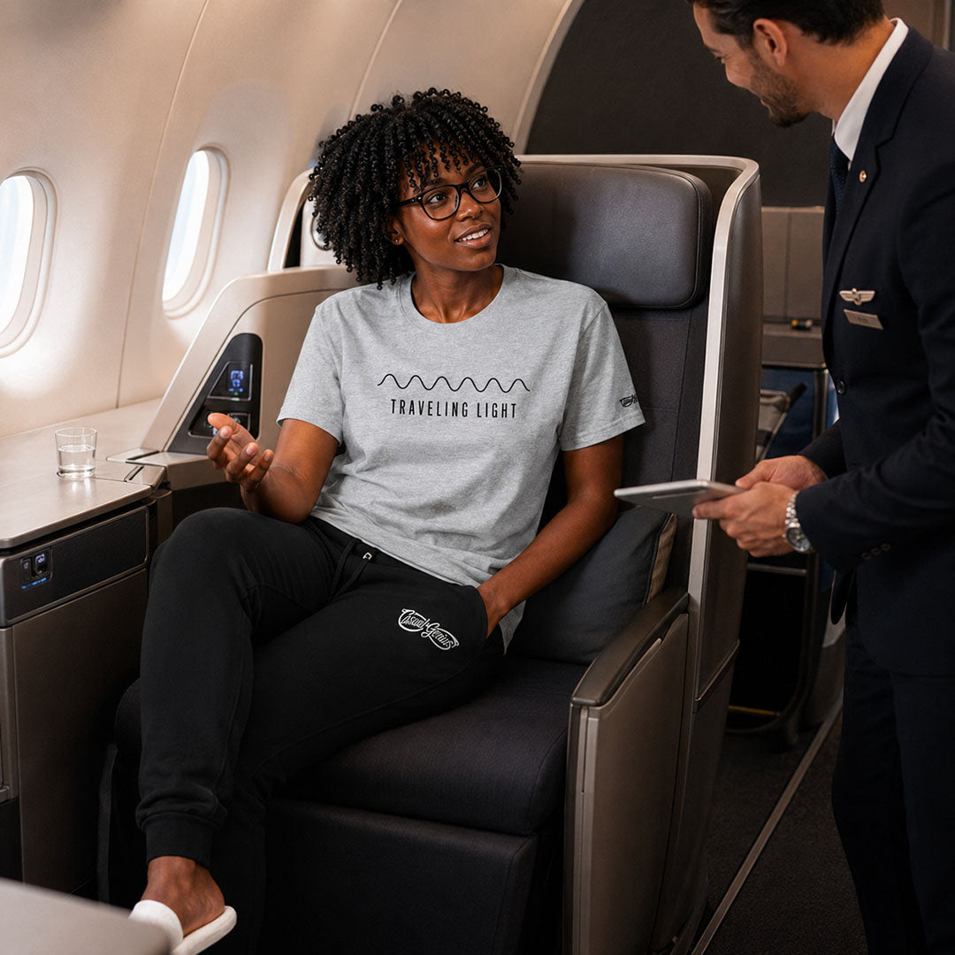 A woman wearing a light gray graphic t-shirt with 'TRAVELING LIGHT' printed on the front, sits in an airplane business class seat while a flight attendant in a navy uniform stands beside her.