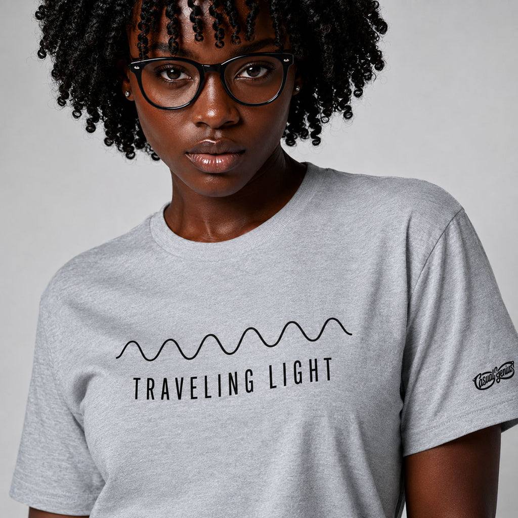 A close-up of a person wearing a light gray heather crewneck t-shirt with a black graphic print that reads 'TRAVELING LIGHT' under a wavy line, set against a plain light gray background.