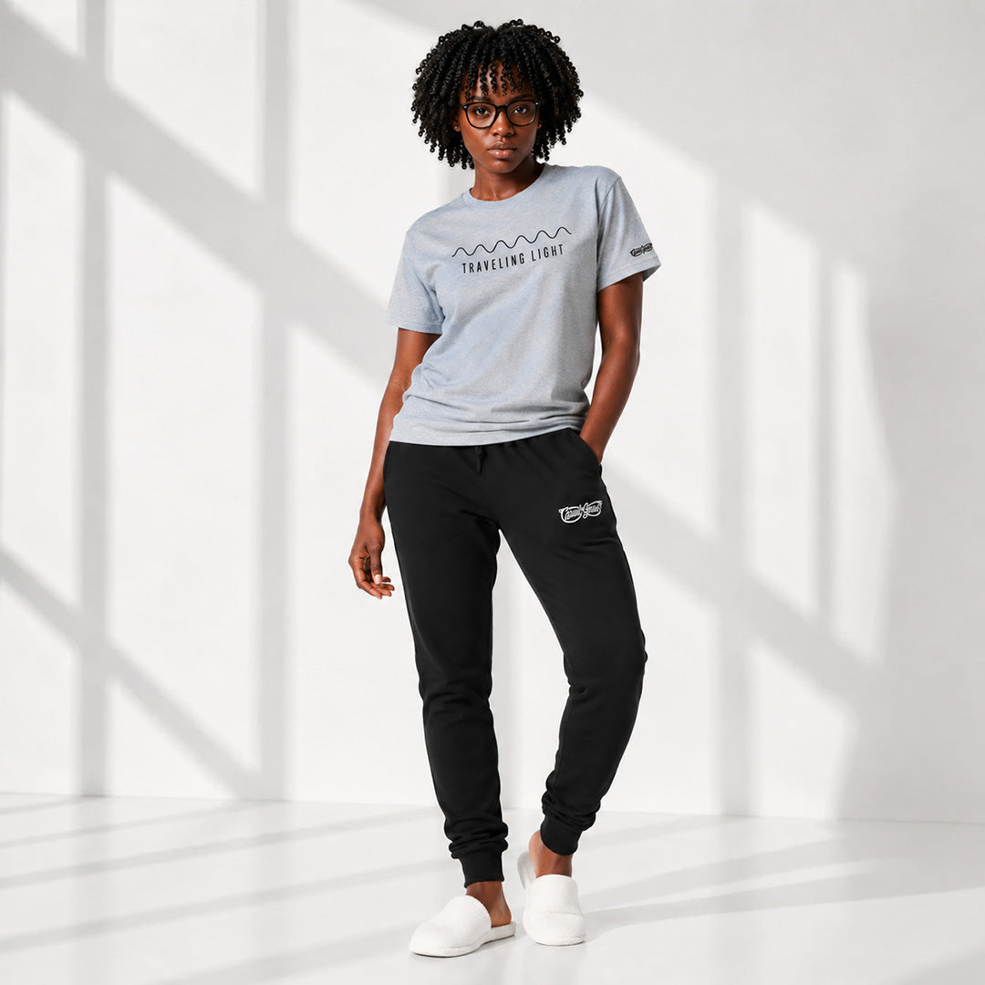 A woman modeling a light gray graphic t-shirt and black jogger pants with white slip-on shoes, standing in a bright studio with window shadow patterns on a white background.