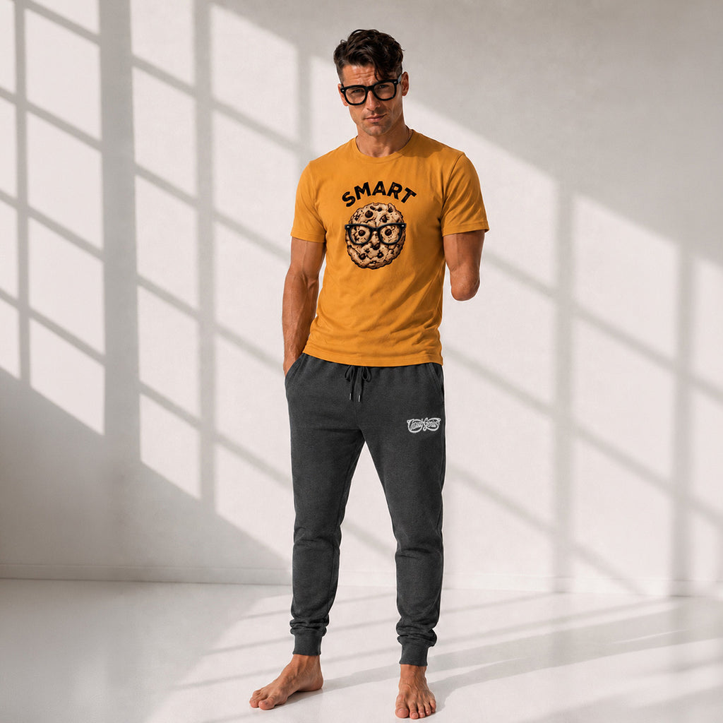 A man wearing a mustard yellow graphic t-shirt and dark gray jogger sweatpants, standing barefoot in a brightly lit room with window shadow patterns on a white wall.